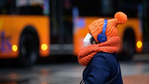 Eine Frau sitzt mit FFP2-Maske, dicker Mütze und Kopfhörern an einem Busbahnhof, während ein Linienbus vorbeifährt., © Jonas Walzberg/dpa